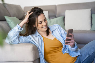 Smiling young woman using mobile phone while sitting at sofa in living room - AFVF08186