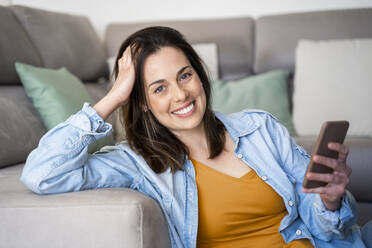 Beautiful smiling woman holding mobile phone while sitting at sofa - AFVF08187