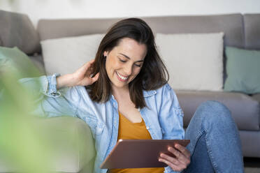 Smiling woman with hand in hair using digital tablet while sitting at sofa in living room - AFVF08188