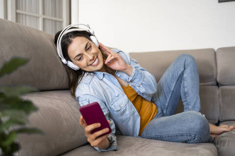 Smiling woman holding mobile phone while listening music through headphones on sofa at home stock photo