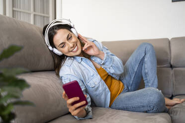 Smiling woman holding mobile phone while listening music through headphones on sofa at home - AFVF08191