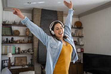 Cheerful woman with headphones enjoying music while standing in living room - AFVF08192