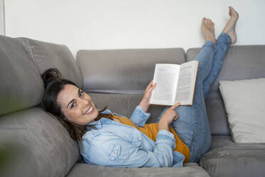 Smiling woman with book lying on sofa - AFVF08194