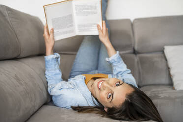 Smiling woman with feet up holding book while lying on sofa at home  - AFVF08195