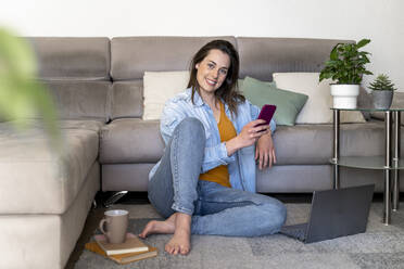Smiling woman with laptop holding mobile phone while sitting at sofa in living room - AFVF08196