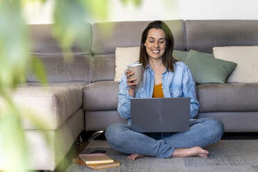 Smiling woman having coffee while using laptop in living room - AFVF08197