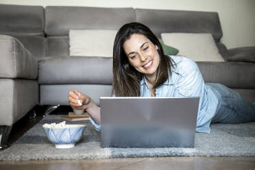 Lächelnde Frau mit Laptop, die Popcorn isst, während sie zu Hause auf dem Teppich liegt - AFVF08201