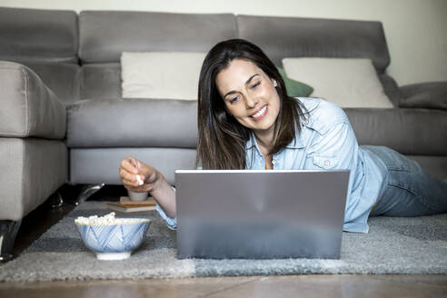 Smiling woman with laptop eating popcorn while lying on carpet at home - AFVF08201