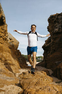 Männlicher Läufer mit ausgestreckten Armen springt über Felsen vor blauem Himmel - JRFF05088