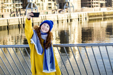 Glückliches Mädchen nimmt Selfie durch Handy am Geländer gegen Wasser - IHF00404