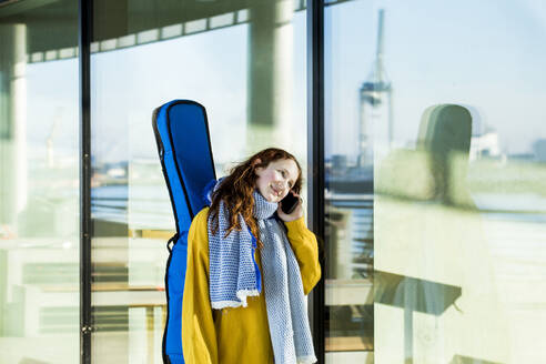 Lächelndes Mädchen, das eine Gitarre trägt, während es mit einem Mobiltelefon an einem Glas spricht - IHF00412