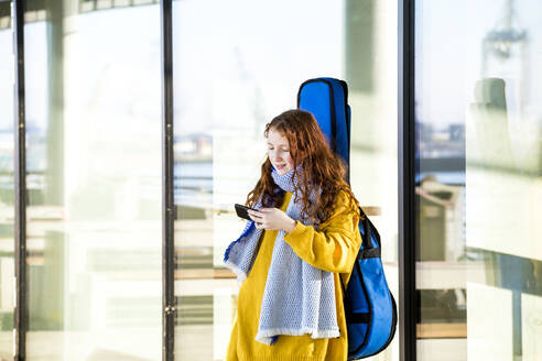 Mädchen mit Gitarre schaut auf Handy, während sie gegen eine Glaswand steht - IHF00413