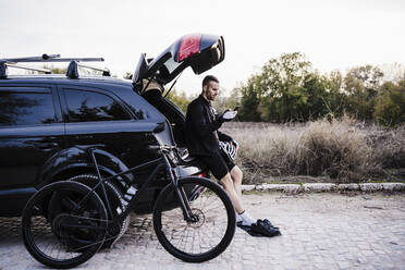 Männlicher Radfahrer, der ein Mobiltelefon benutzt, während er mit dem Fahrrad im Kofferraum eines Autos sitzt - EBBF02511