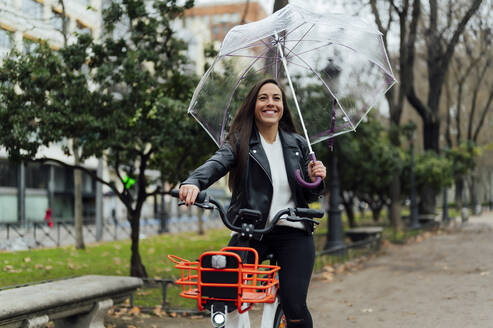 Schöne Frau fährt Elektrofahrrad und hält Regenschirm auf der Straße in der Stadt - PGF00457
