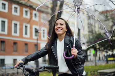Woman holding umbrella while riding bicycle at street in city - PGF00458