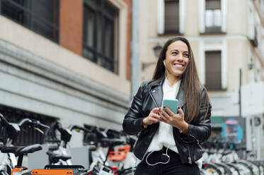 Lächelnde Frau hört Musik durch Smartphone auf der Straße in der Stadt - PGF00459