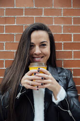 Smiling woman in leather jacket holding reusable cup against brick wall - PGF00461