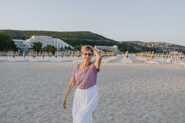 Ältere Frau mit Hand in den Haaren steht gegen einen Hügel am Strand - OGF00885