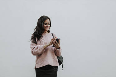 Smiling woman using smart phone while standing against white wall - GMLF00976