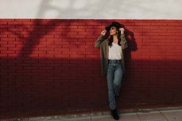Schöne Frau in Strickjacke und Hut lehnt sich an eine rote Backsteinmauer und schaut weg - GMLF00980