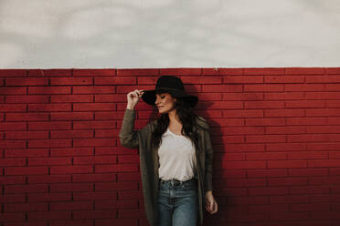 Woman holding hat with eyes closed standing against red brick wall - GMLF00981