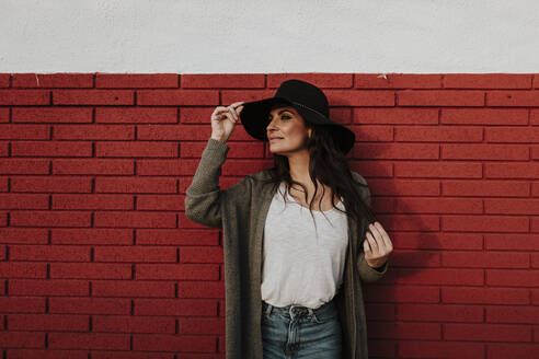 Frau mit Hut schaut weg, während sie an einer roten Backsteinmauer steht - GMLF00982