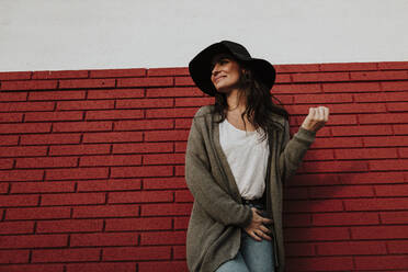 Smiling woman in cardigan looking away while standing against red brick wall - GMLF00983