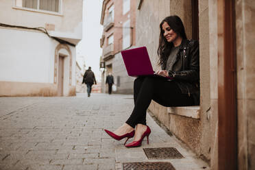 Businesswoman in red stilettos sitting on doorway while using laptop - GMLF00984