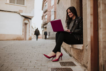 Geschäftsfrau in roten Stilettos, die an der Tür sitzt und einen Laptop benutzt - GMLF00984