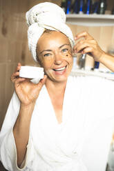 Smiling mature woman applying moisturizer to face in bathroom - PSIF00440