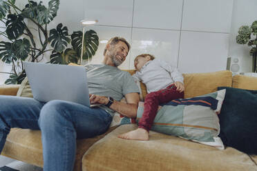 Happy father with laptop and son on sofa in living room - MFF07156