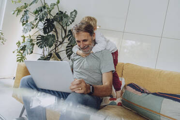 Father with laptop and playful son on sofa in living room - MFF07157