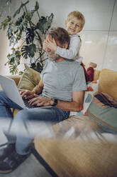 Smiling playful boy covering father's eye on sofa in living room - MFF07159