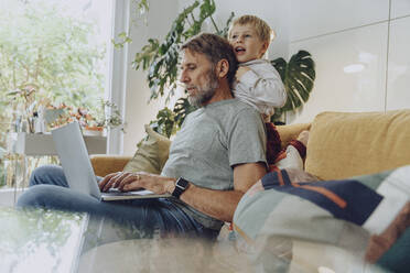 Playful boy leaning on father using laptop at home - MFF07160