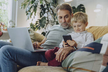 Vater schaut auf den Jungen, der sein Smartphone benutzt, während er zu Hause auf dem Sofa sitzt - MFF07161
