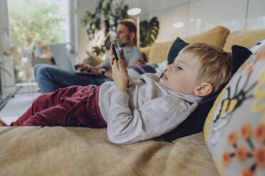 Boy using phone while lying on sofa in living room - MFF07163