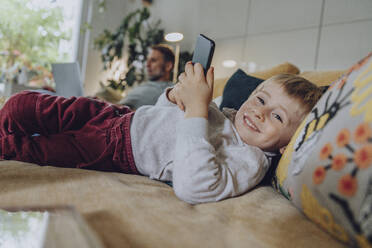 Lächelnder Junge, der ein Mobiltelefon benutzt, während er zu Hause auf dem Sofa liegt - MFF07164