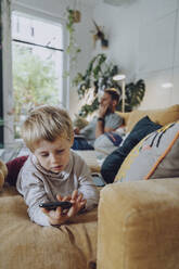 Boy using mobile phone while lying on sofa in living room - MFF07165