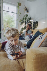 Junge, der ein Mobiltelefon benutzt, während er auf dem Sofa im Wohnzimmer liegt - MFF07165