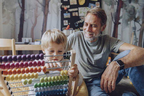 Happy father watching son counting abacus in bedroom - MFF07174