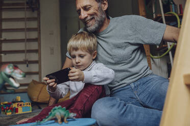 Father watching son playing games on smart phone at home - MFF07180