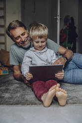 Lächelnder Vater beobachtet seinen Sohn bei der Nutzung eines Tablets im Schlafzimmer - MFF07181