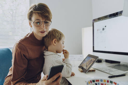 Eine berufstätige Mutter benutzt ein Smartphone, während ihr Sohn ein Video auf einem digitalen Tablet ansieht - MFF07187