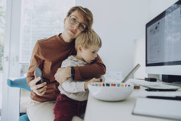 Liebevolle Mutter, die ihren Sohn umarmt, während sie im Büro zu Hause auf ein digitales Tablet schaut - MFF07188