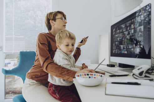 Mother using computer while son sitting on her lap at home office - MFF07190