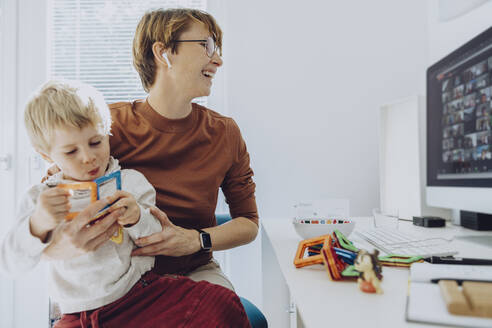 Mid adult businesswoman working while son sitting on her lap playing with toys at home office - MFF07193