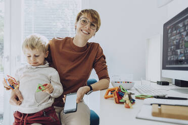 Lächelnde Mutter mit Sohn, der auf dem Schoß sitzt und mit Spielzeug im Büro zu Hause spielt - MFF07195