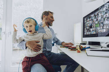 Geschäftsmann bei der Arbeit, während sein Sohn mit kabellosen Kopfhörern auf dem Schoß im Heimbüro sitzt - MFF07197