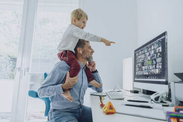 Älterer Geschäftsmann, der an einer Videokonferenz teilnimmt, während sein Sohn auf seiner Schulter sitzt und auf den Computerbildschirm im Heimbüro zeigt - MFF07202