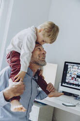 Son kissing father's forehead while sitting on his shoulder at home office - MFF07205